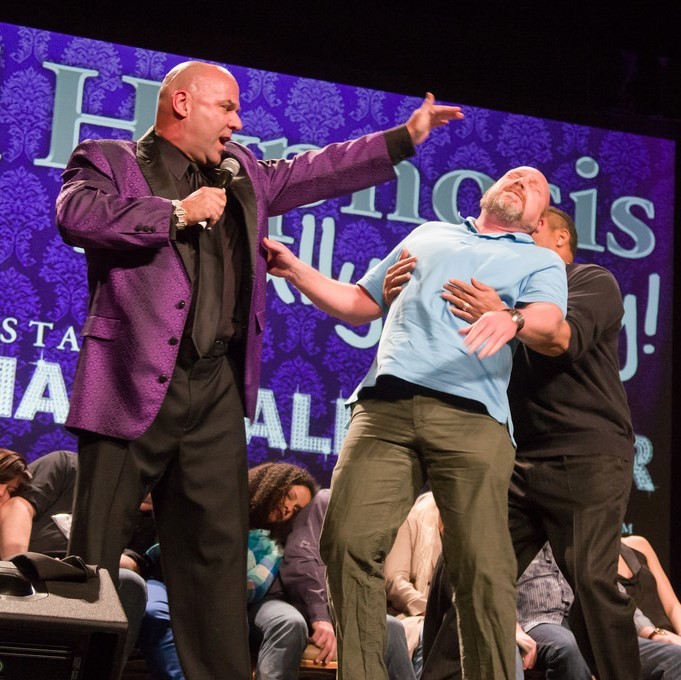 A stage hypnotist holds his hand over the head of a volunteer who falls limp into the arms of the hypnotist's assistant. A group of volunteers seem to be unconscious in their seats in the background.