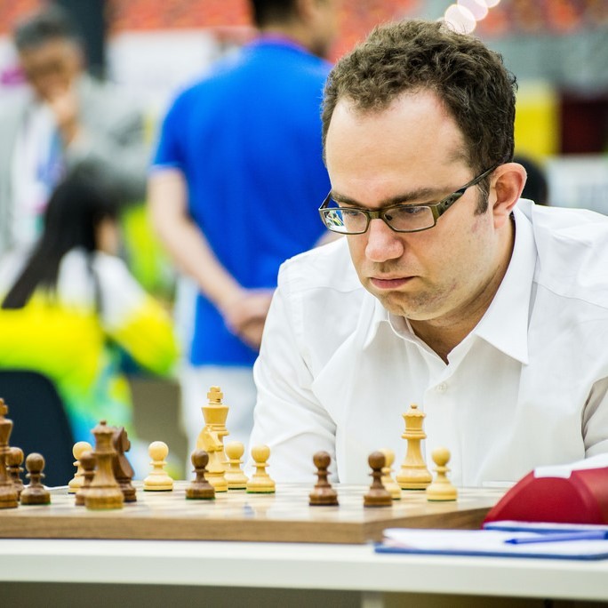 A man sits hunched over looking at the pieces on a chessboard with an expression of deep concentration on his face.